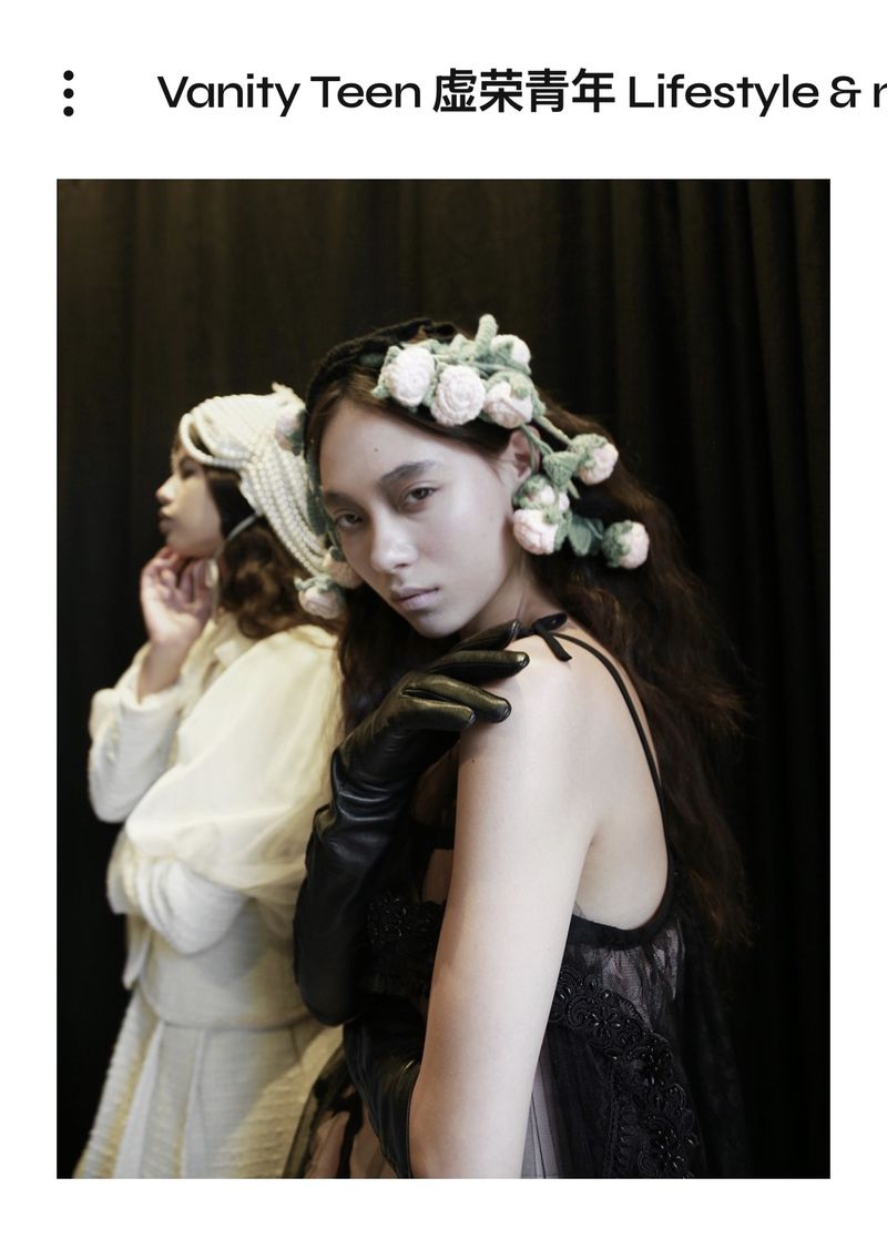 Two women pose in an indoor setting with a dark background, one wearing a floral headpiece and black gloves, the other in white.