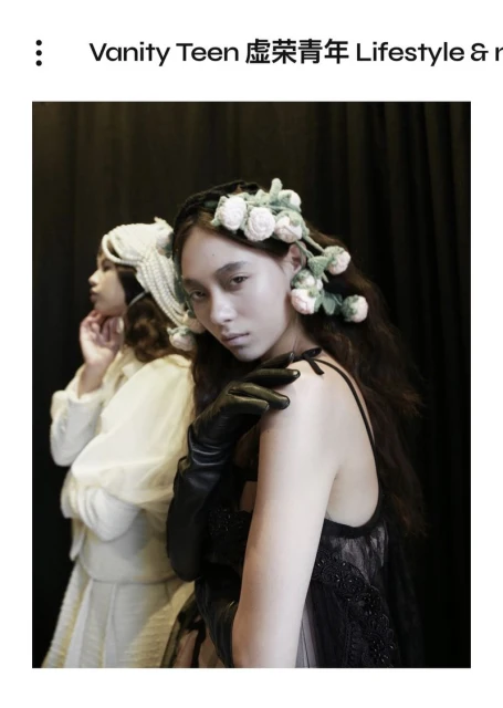 Two women pose in an indoor setting with a dark background, one wearing a floral headpiece and black gloves, the other in white.