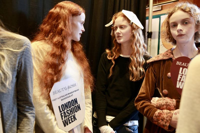 Backstage shot featuring three red-haired fashion models at London Fashion Week, holding promotional signs, medium shot.