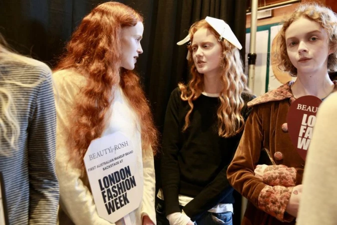 Backstage shot featuring three red-haired fashion models at London Fashion Week, holding promotional signs, medium shot.