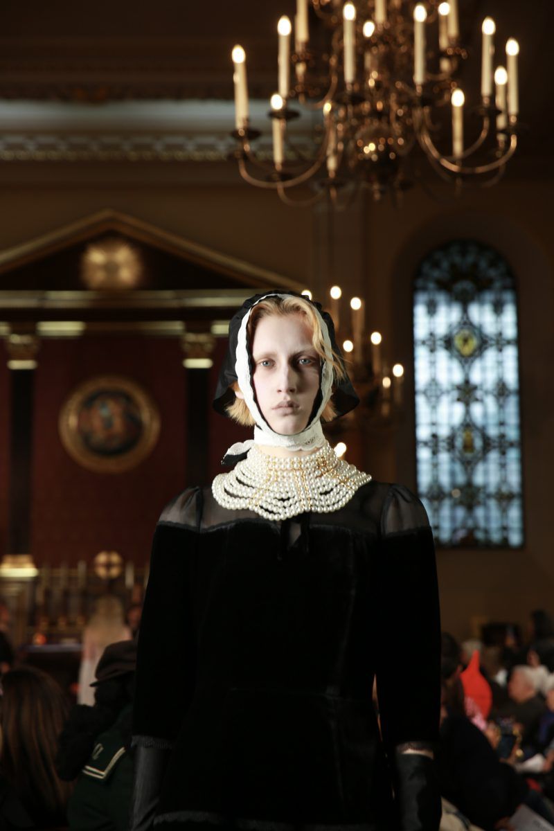Adult female runway model showcases dark dress with pearl collar in an indoor setting with chandeliers and seated onlookers in the background.