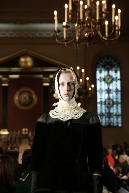 Adult female runway model showcases dark dress with pearl collar in an indoor setting with chandeliers and seated onlookers in the background.