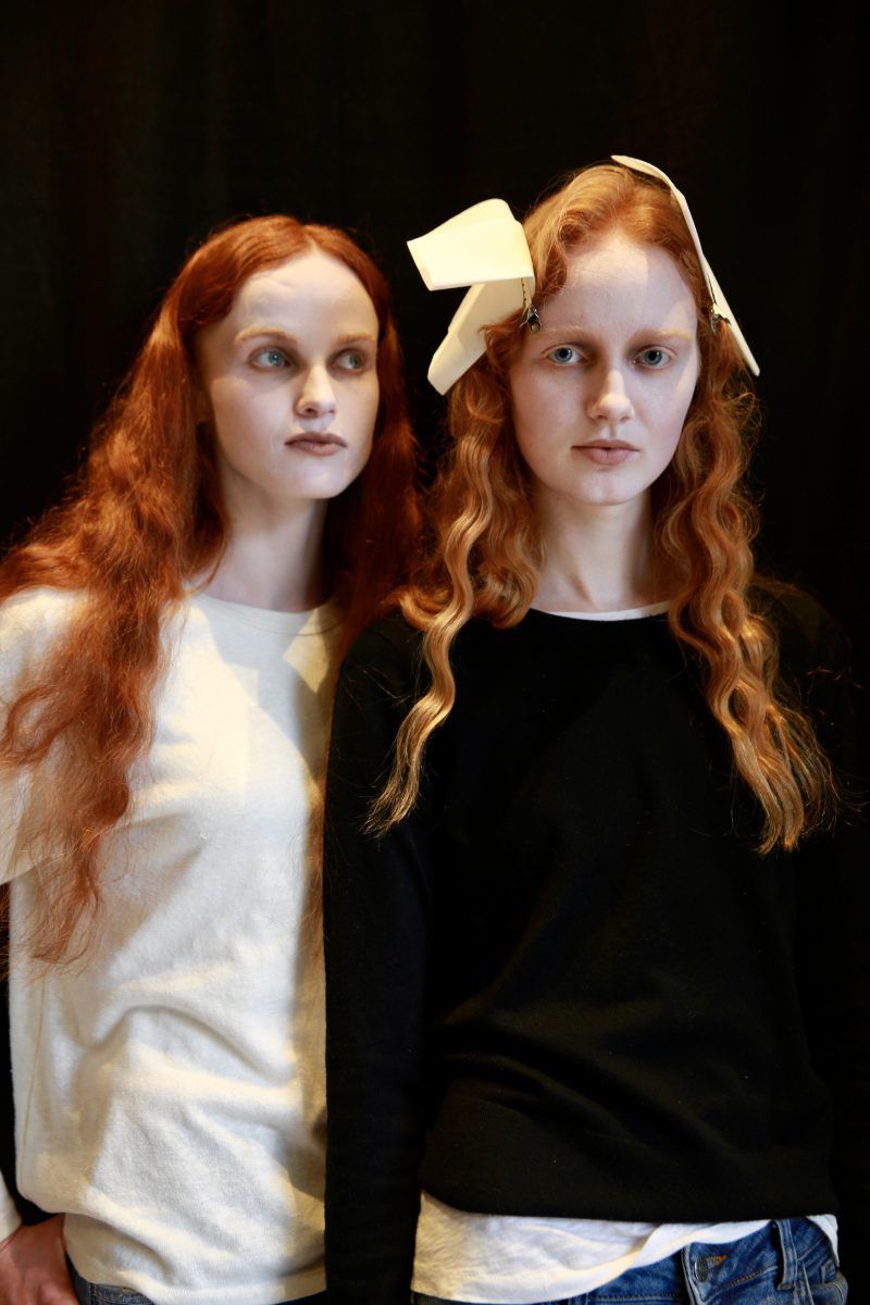 Two models with red hair wearing black and white tops pose in front of a dark background. One has white paper in her hair.