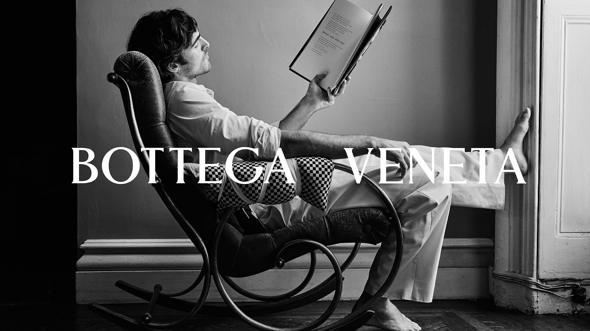 Monochrome shot of a man in a rocking chair reading a book, in a relaxed pose in an indoor setting, with the 'Bottega Veneta' logo overlayed.
