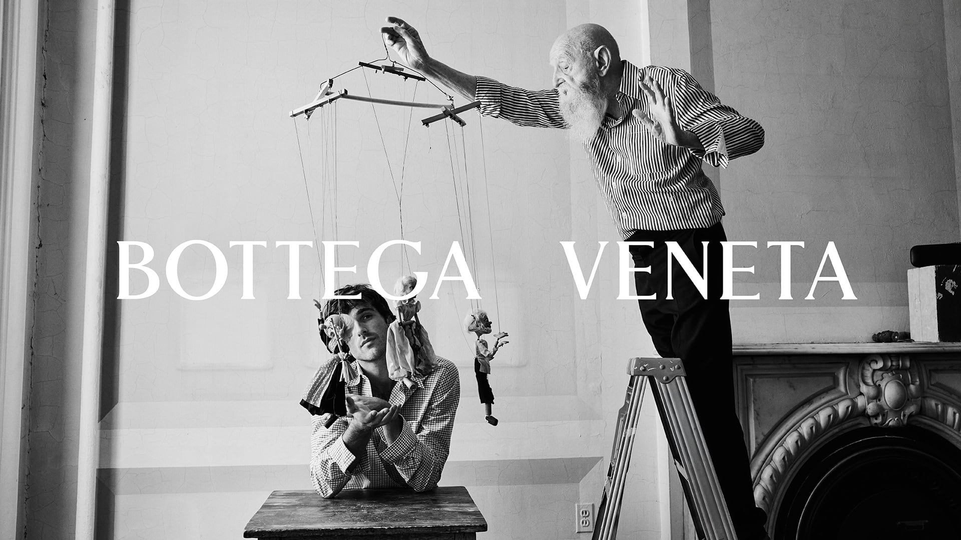 Black-and-white image features an elderly man on a ladder operating marionette puppets, with a younger man and vintage fireplace.