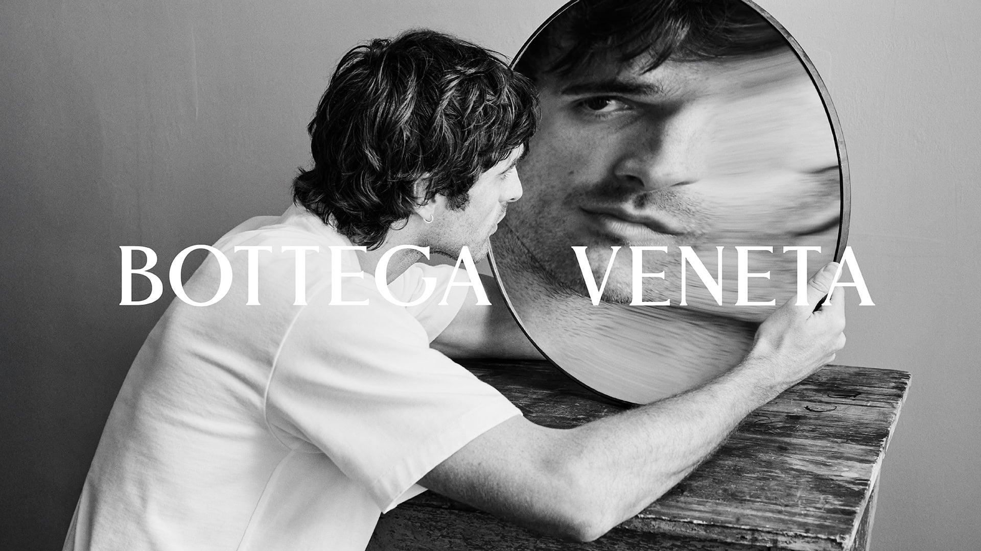 Black and white portrait of a man looking at his reflection in a round mirror on a wooden surface. Brand name BOTTEGA VENETA visible.
