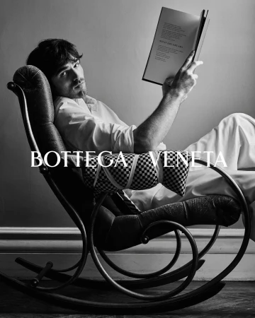 Black and white fashion shot of a dark-haired man relaxing in a rocking chair with a book in a bright, naturally lit indoor setting.