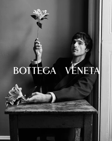 Jacob Elordi in a dark suit holding a white lily at a wooden table in a monochrome portrait for Bottega Veneta Fall 2025 campaign