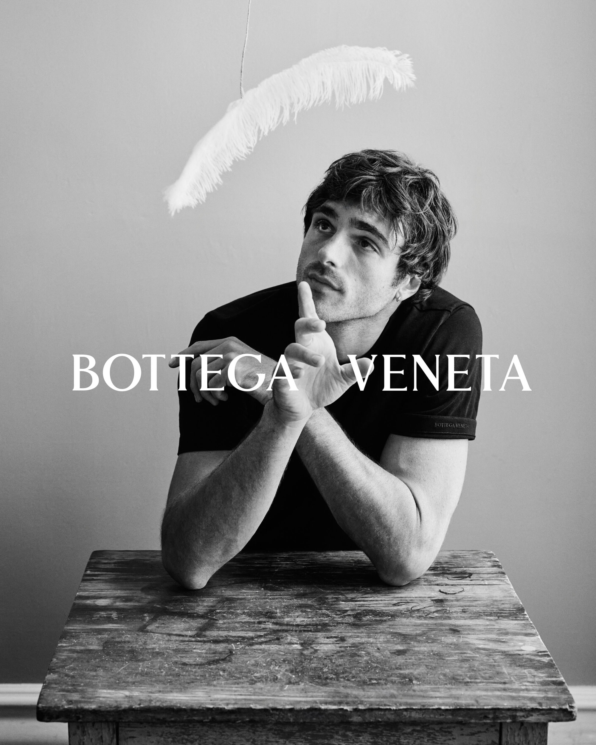 Monochrome portrait of a man with short hair wearing a t-shirt sitting at a wooden table with feather and the text BOTTEGA VENETA.