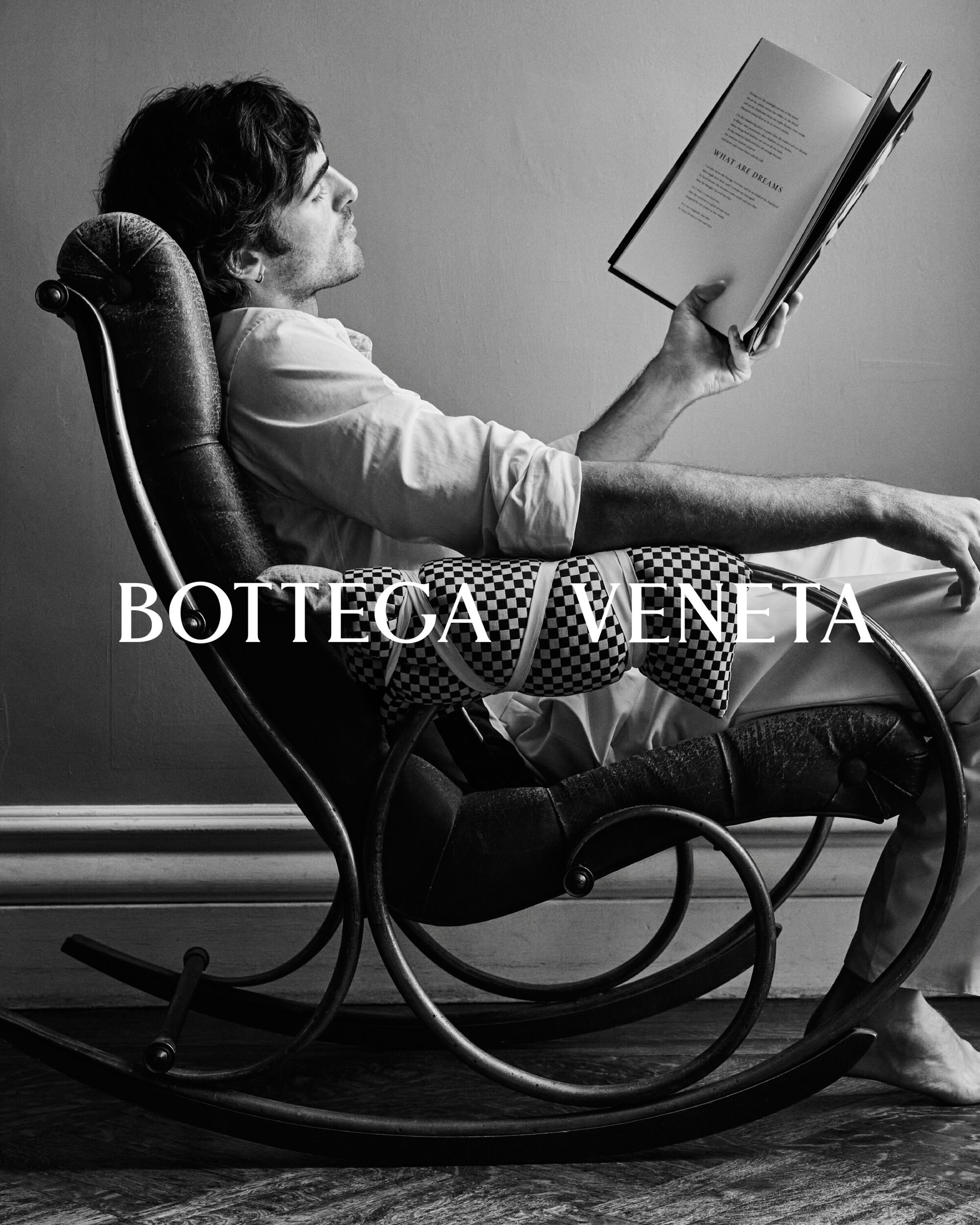 Black and white shot of a man in a rocking chair reading a book. He wears a white shirt and is barefoot. Bottega Veneta text overlay.