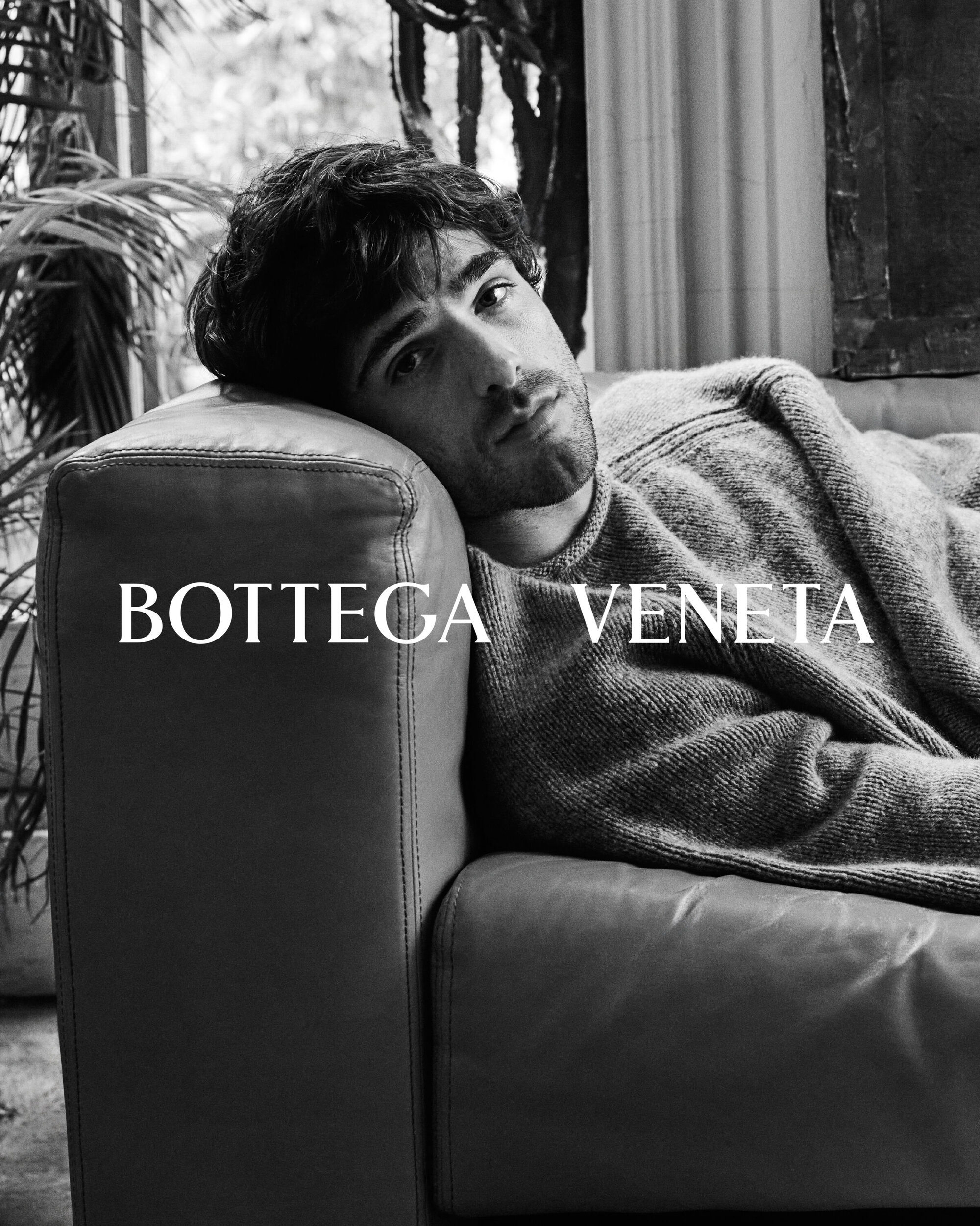 Black and white portrait of a male model reclining on a leather sofa in front of indoor plants for Bottega Veneta advertisement.