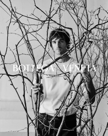 Monochrome portrait of a man standing among bare branches, wearing a white t-shirt and jeans, with Bottega Veneta text overlay.