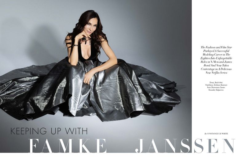 Famke Janssen poses in a silver dress. She is seated with her hand near her face with text overlay on a grey gradient background.