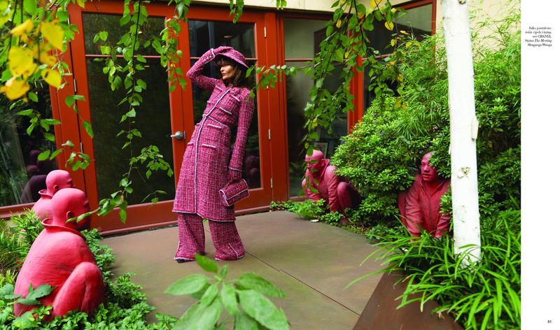 Fashion editorial shot outdoors featuring a woman in a pink tweed suit and matching hat, posing amongst garden foliage and red sculptures.