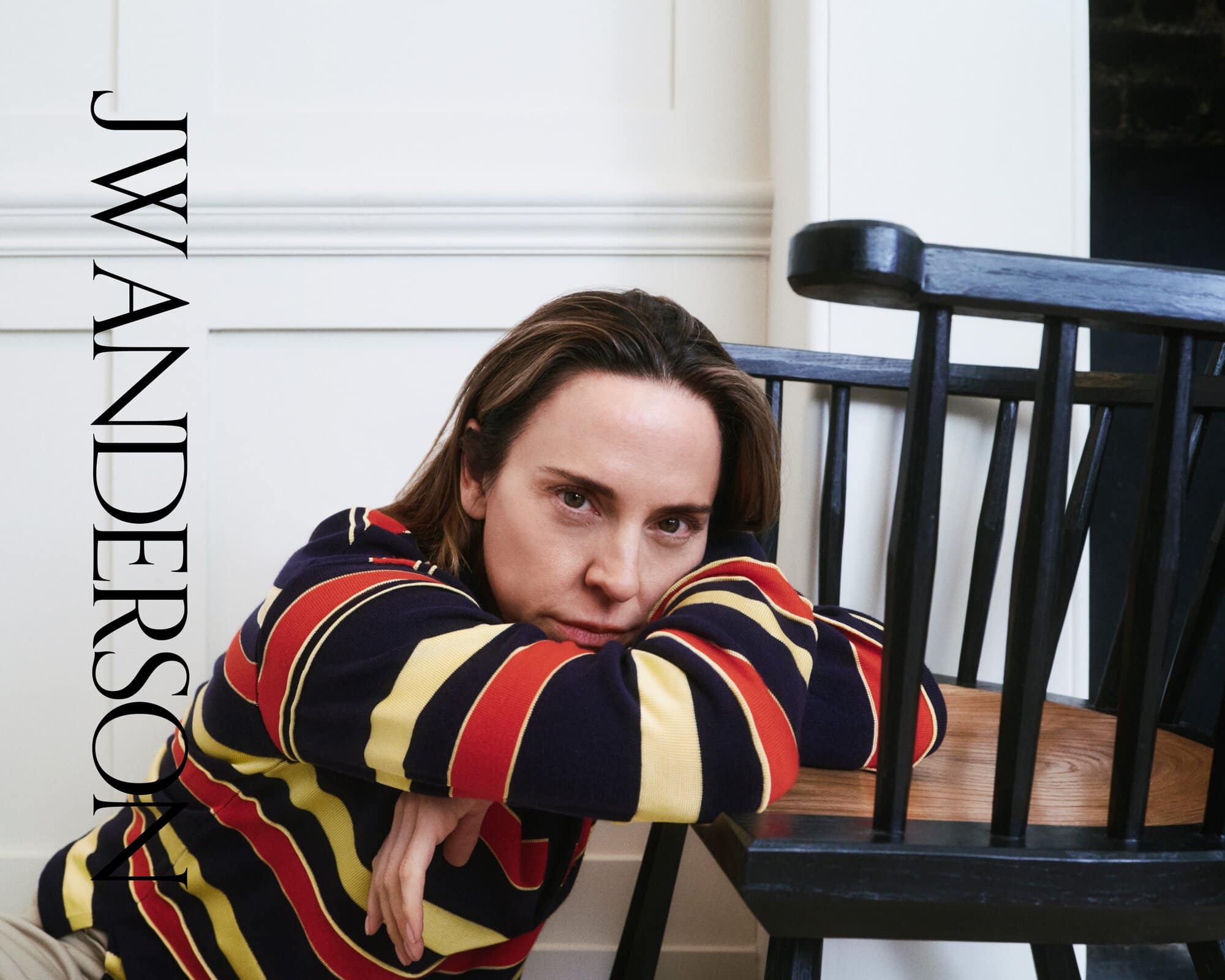 Portrait of a young adult woman with shoulder-length brown hair in a colorful striped sweater, leaning on a black spindle chair.