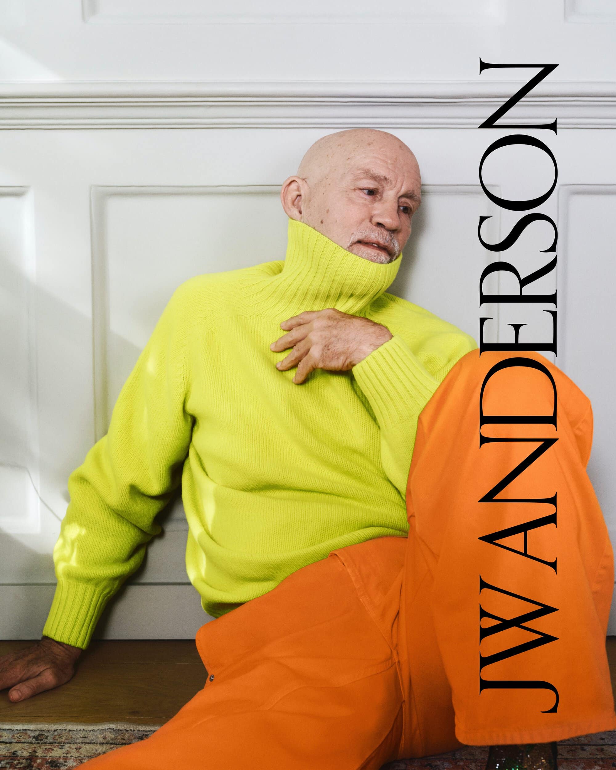 Elderly male model in a bright yellow turtleneck and orange pants poses indoors against a white wall with vertical text overlay.