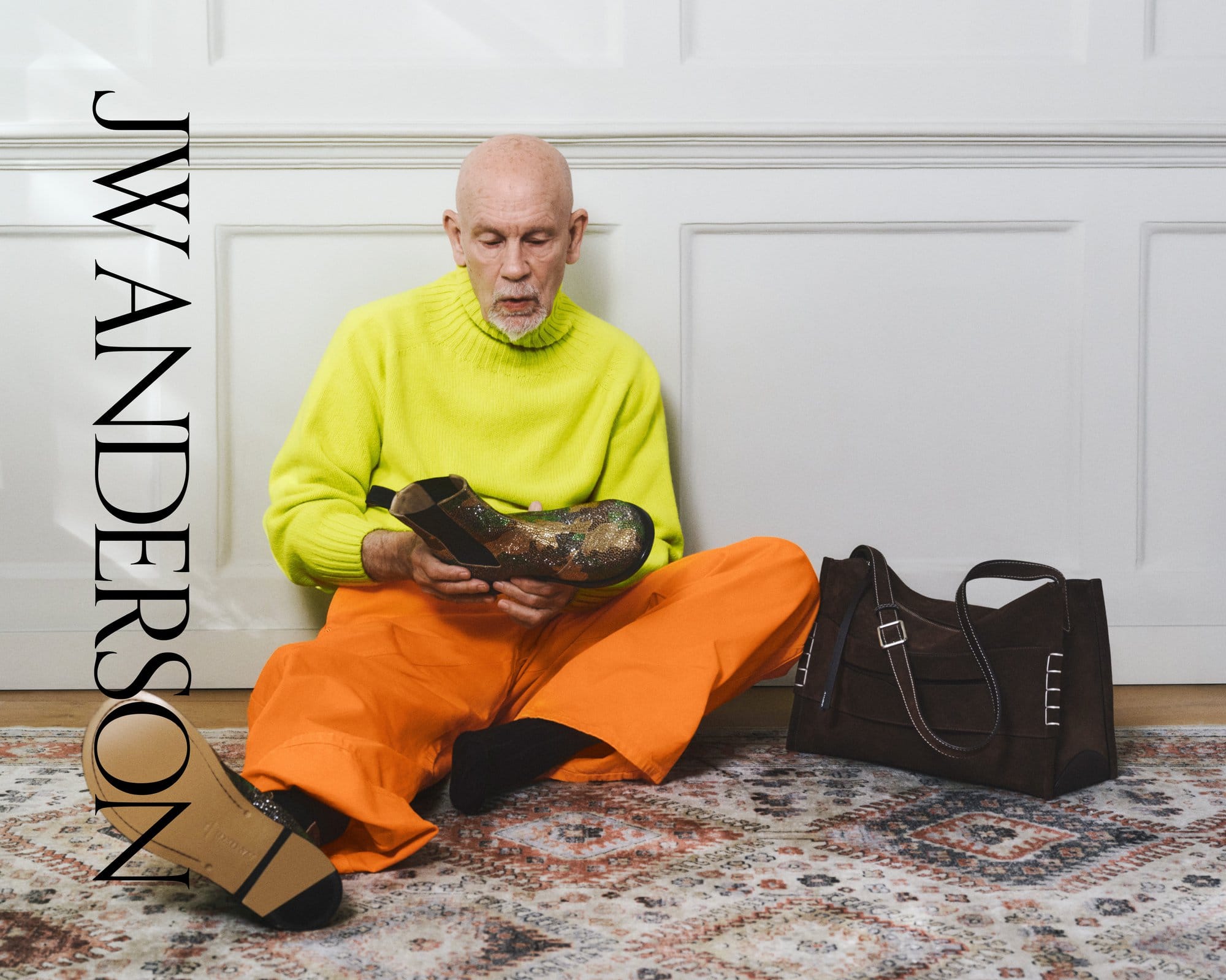 Older man in a neon turtleneck and orange pants sits on the floor, carefully holding a designer ankle boot, styled indoors.
