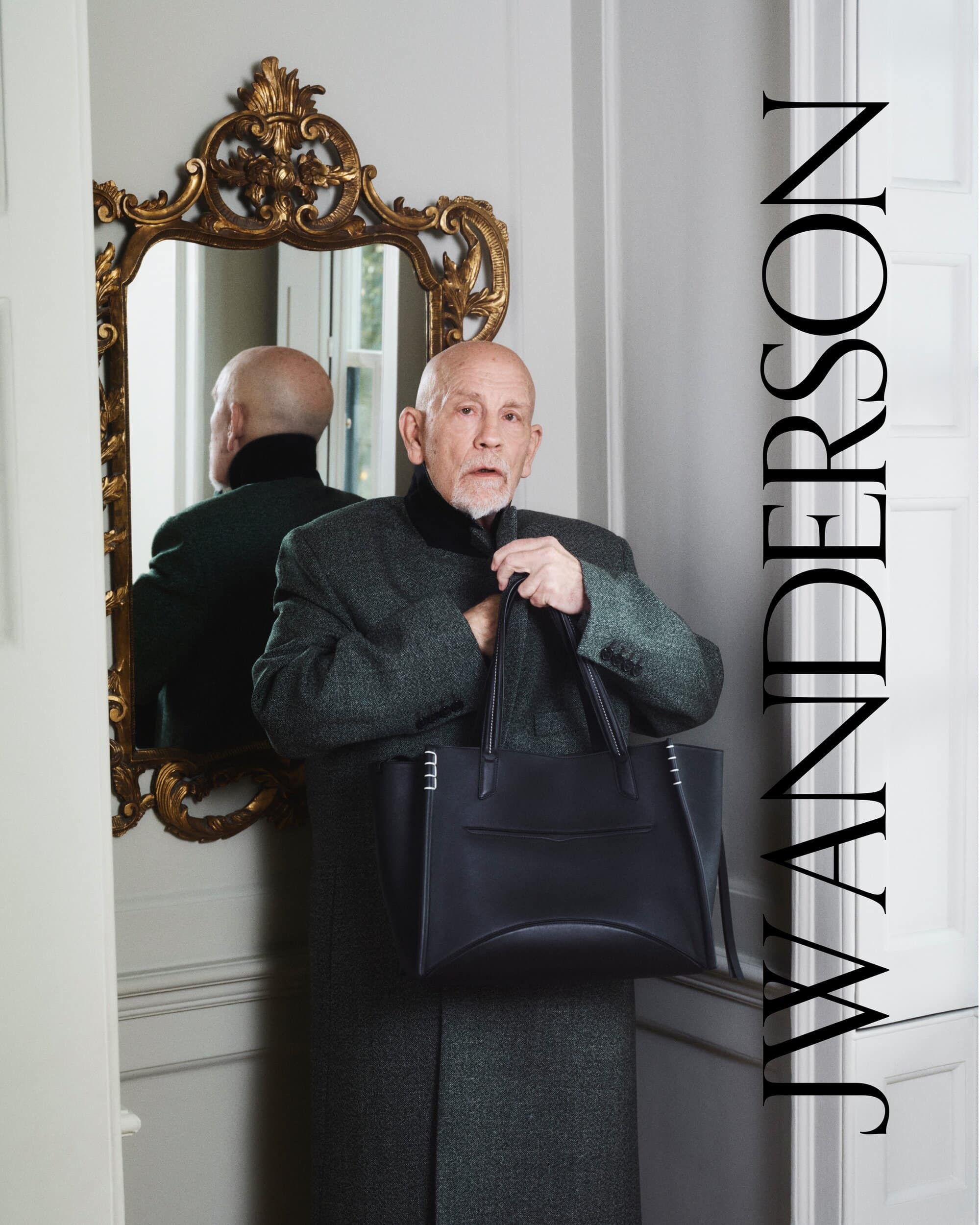 Mature man in a coat and tie holds a black leather tote bag indoors with a gold-framed mirror, posing for J.W. Anderson fashion brand.