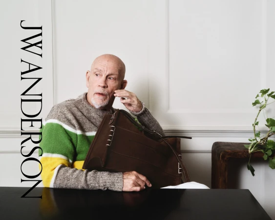 Elegant portrait of a mature male model sitting at a table, holding a brown leather bag, wearing a striped sweater in an indoor setting.