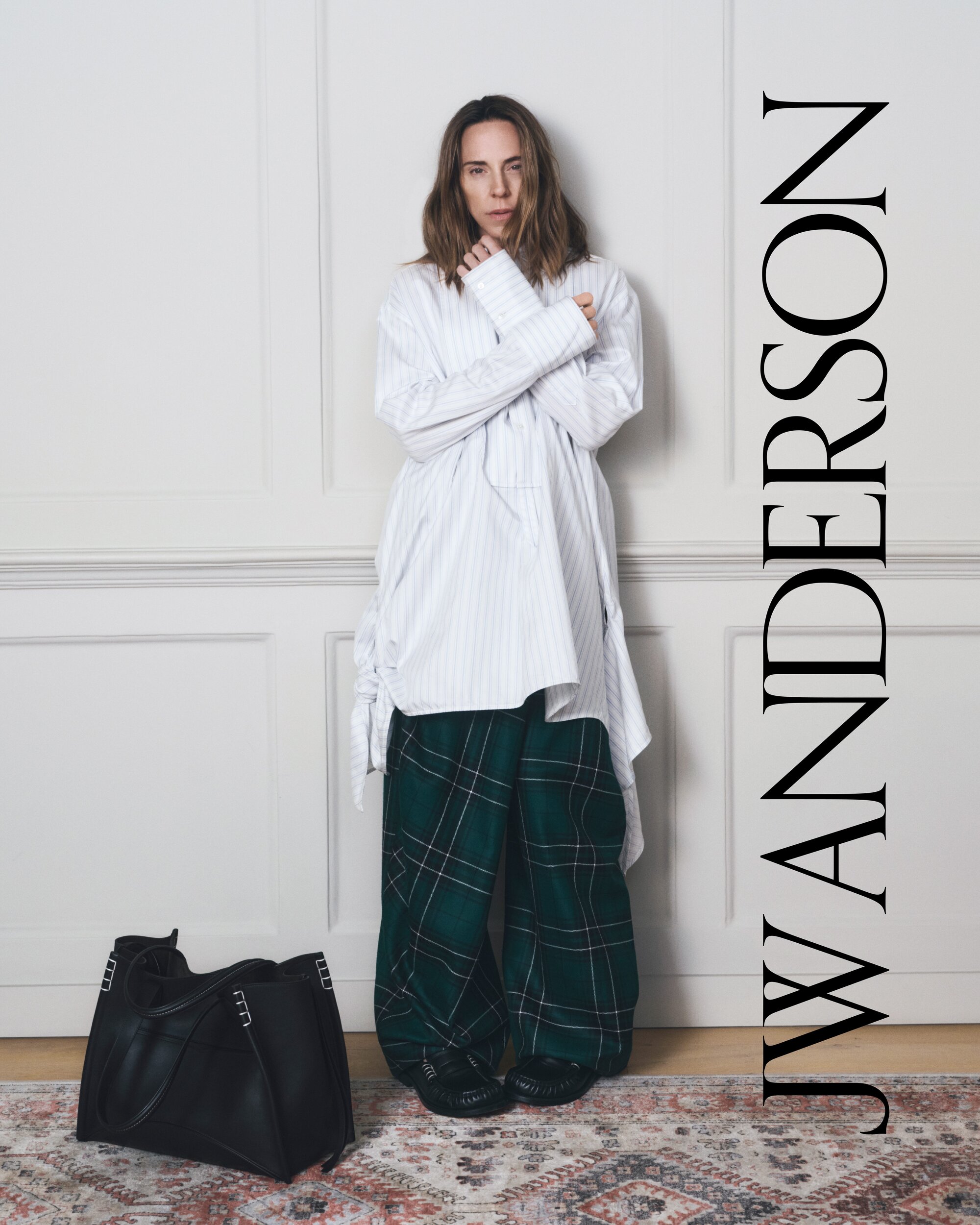 Adult female wearing oversized shirt and plaid pants poses in front of a white wall. She is standing with a black purse.