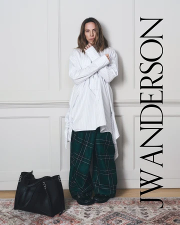 Adult female wearing oversized shirt and plaid pants poses in front of a white wall. She is standing with a black purse.