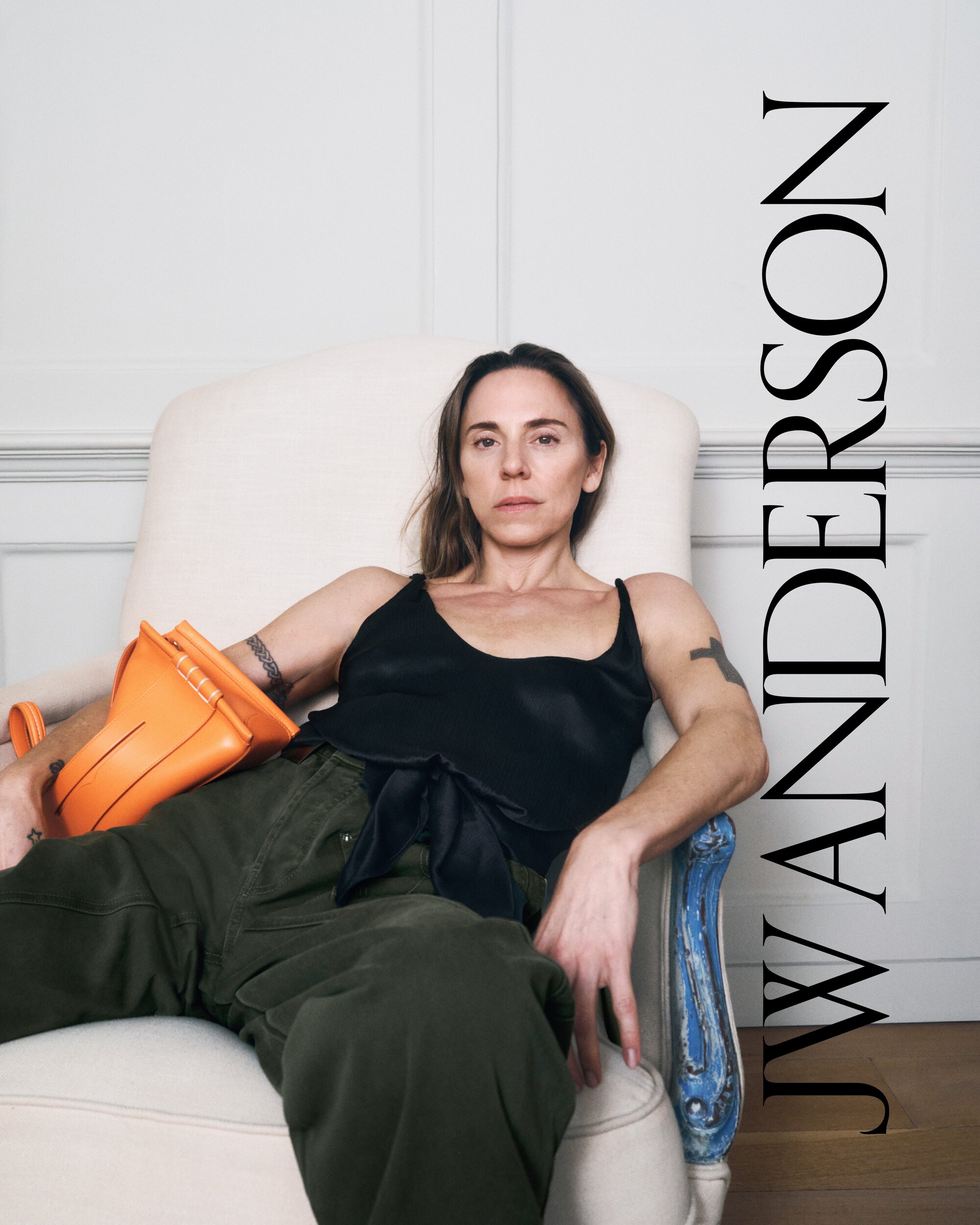 Portrait of Mel C sitting in chair holding orange JW Anderson bag, wearing a black tank top and green pants in a light-filled interior.