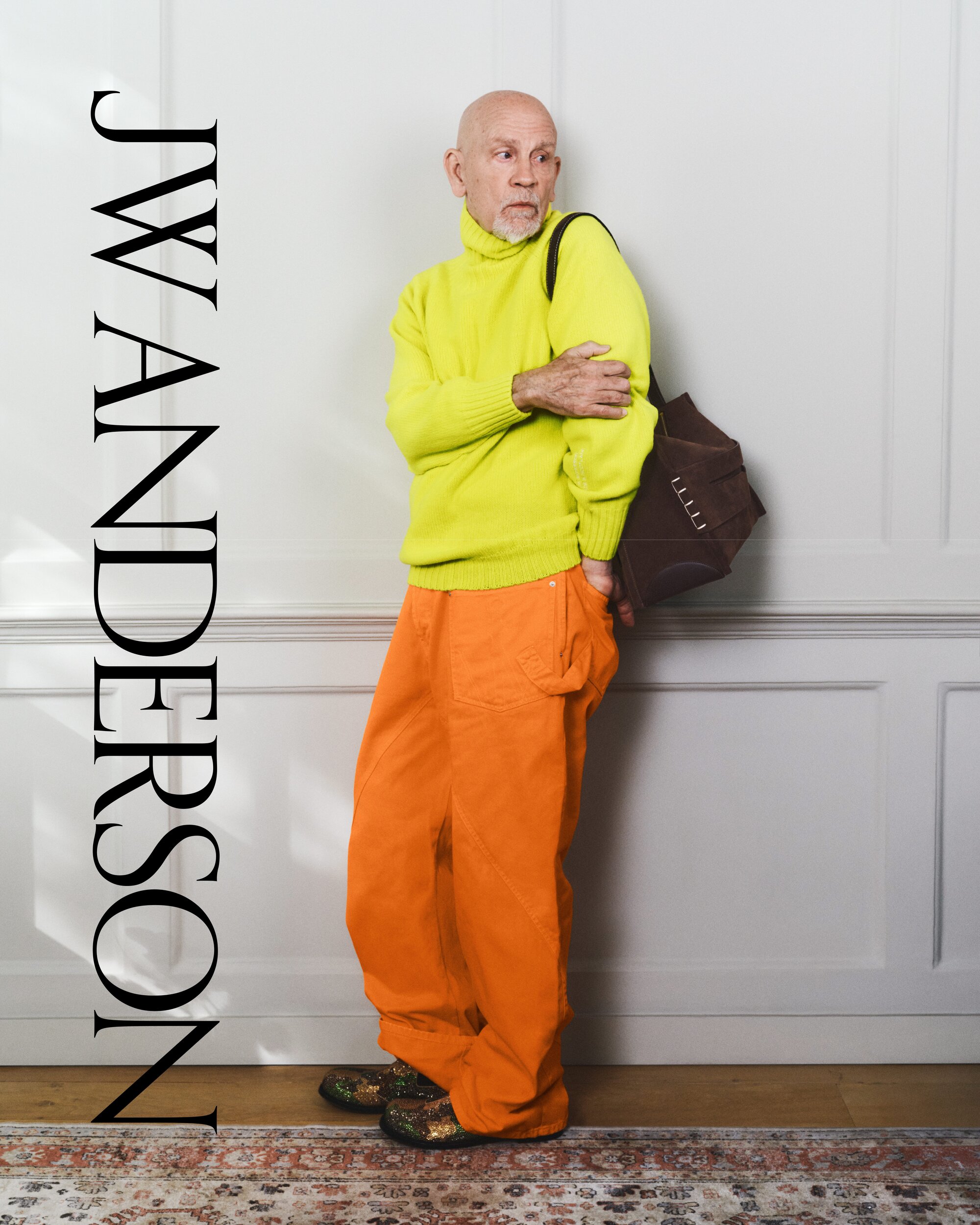 Older adult male posing indoors, wearing a yellow sweater, orange pants, brown bag, and patterned shoes with white wall background.