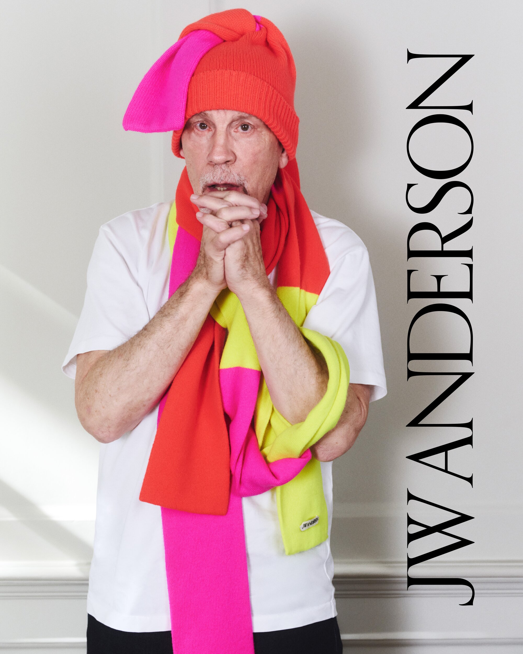 Adult male wearing a bright pink and orange hat and colorful scarf in a studio portrait with a white wall as the backdrop.