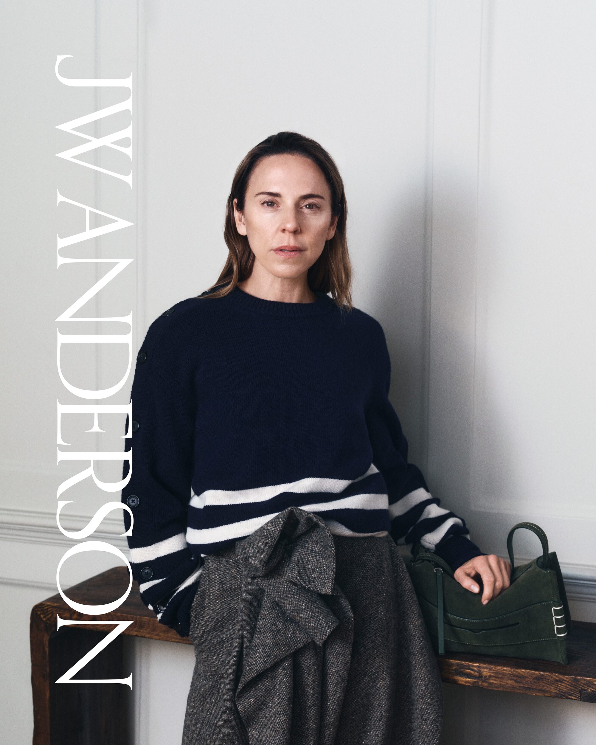 A woman in a navy and white striped sweater poses indoors, holding a dark green handbag. She stands near a wooden console table.
