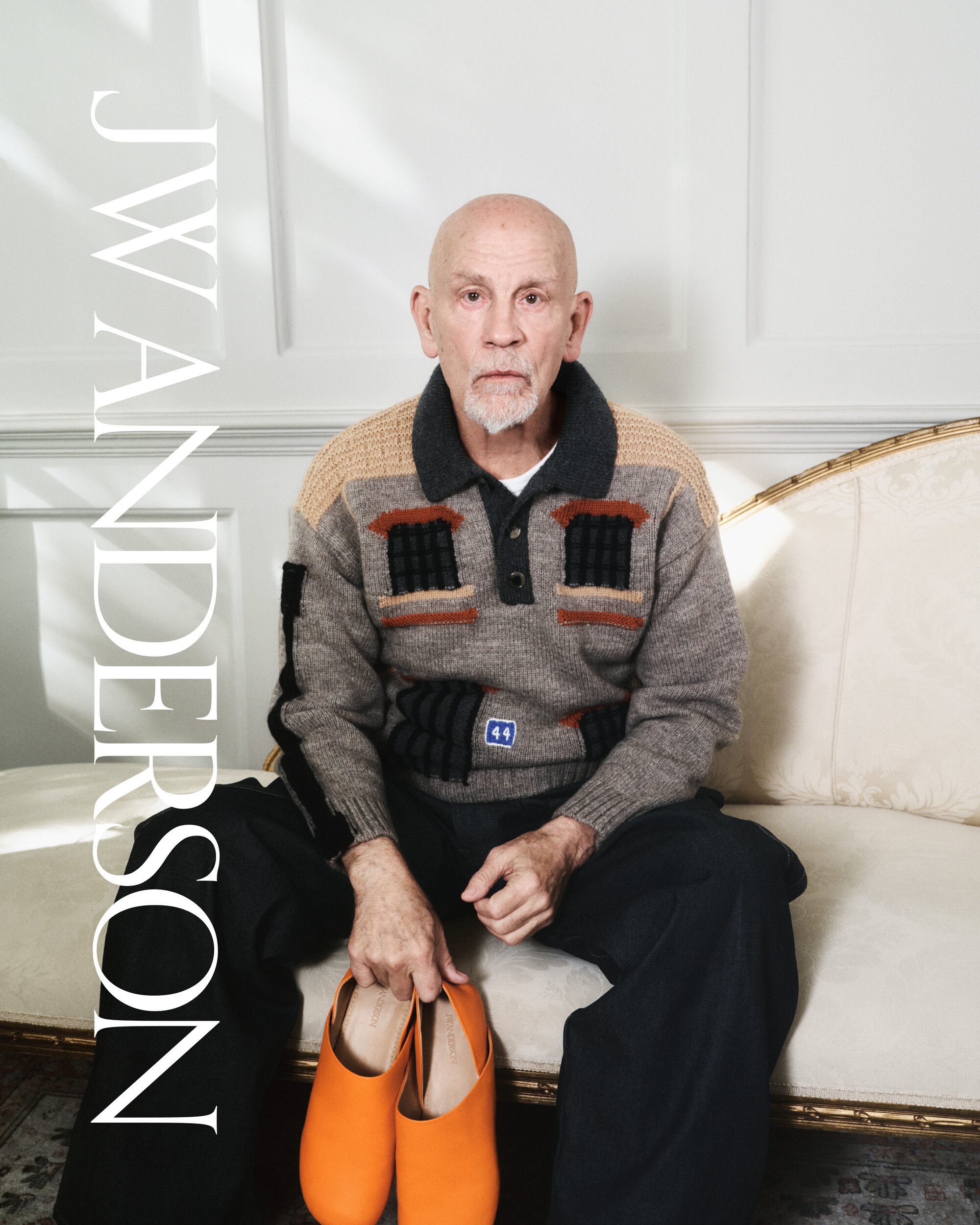 A man sits indoors on an ornate couch, holding bright orange clogs in his hands, wearing a patterned sweater and dark pants.