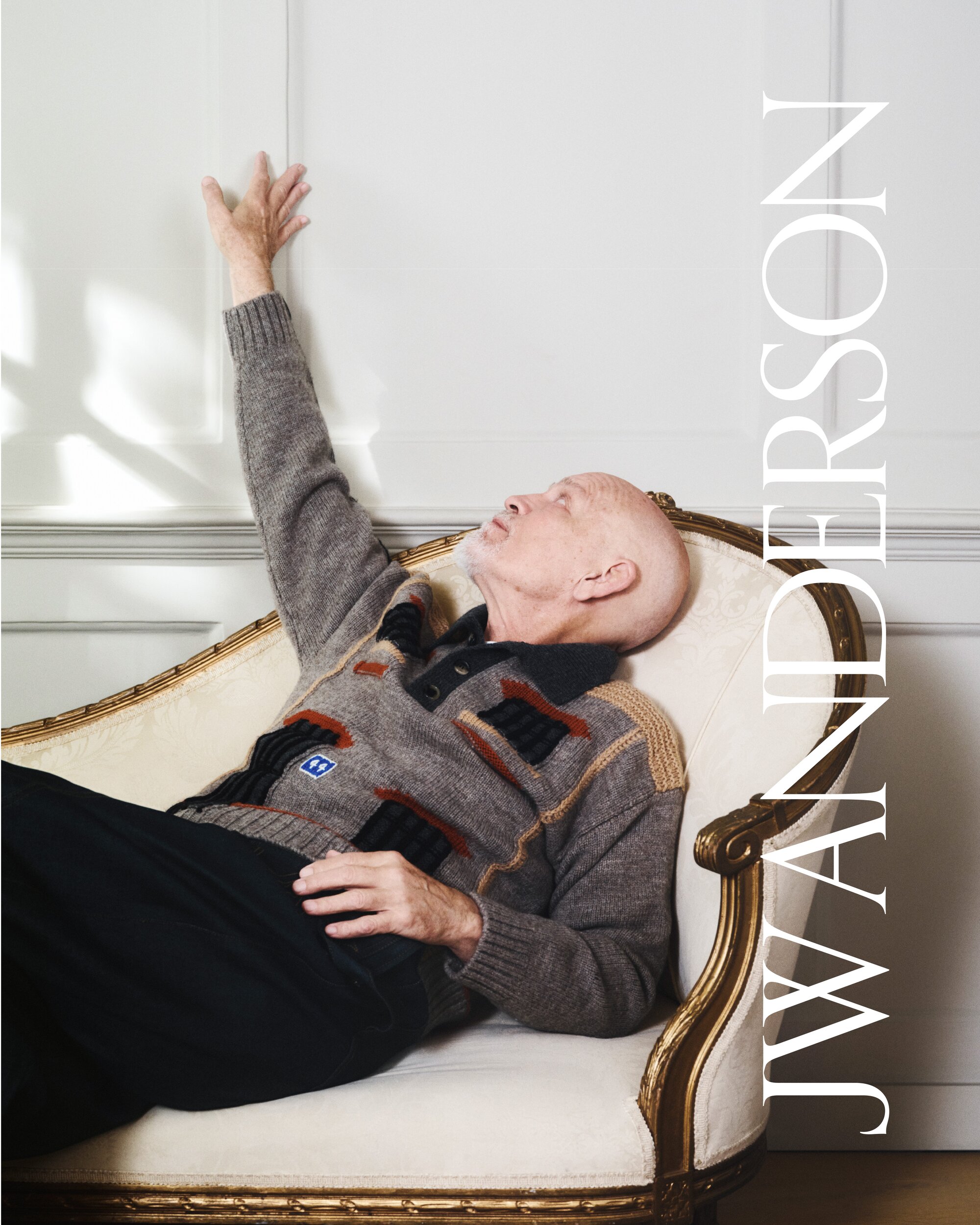 A bald adult male in a patterned sweater reclines on an ornate white and gold couch with his arm reaching up towards a white wall.