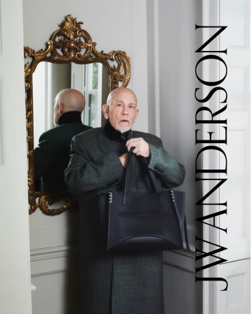 John Malkovich holding a dark leather tote bag wearing a textured grey coat standing beside an ornate mirror in a hotel room