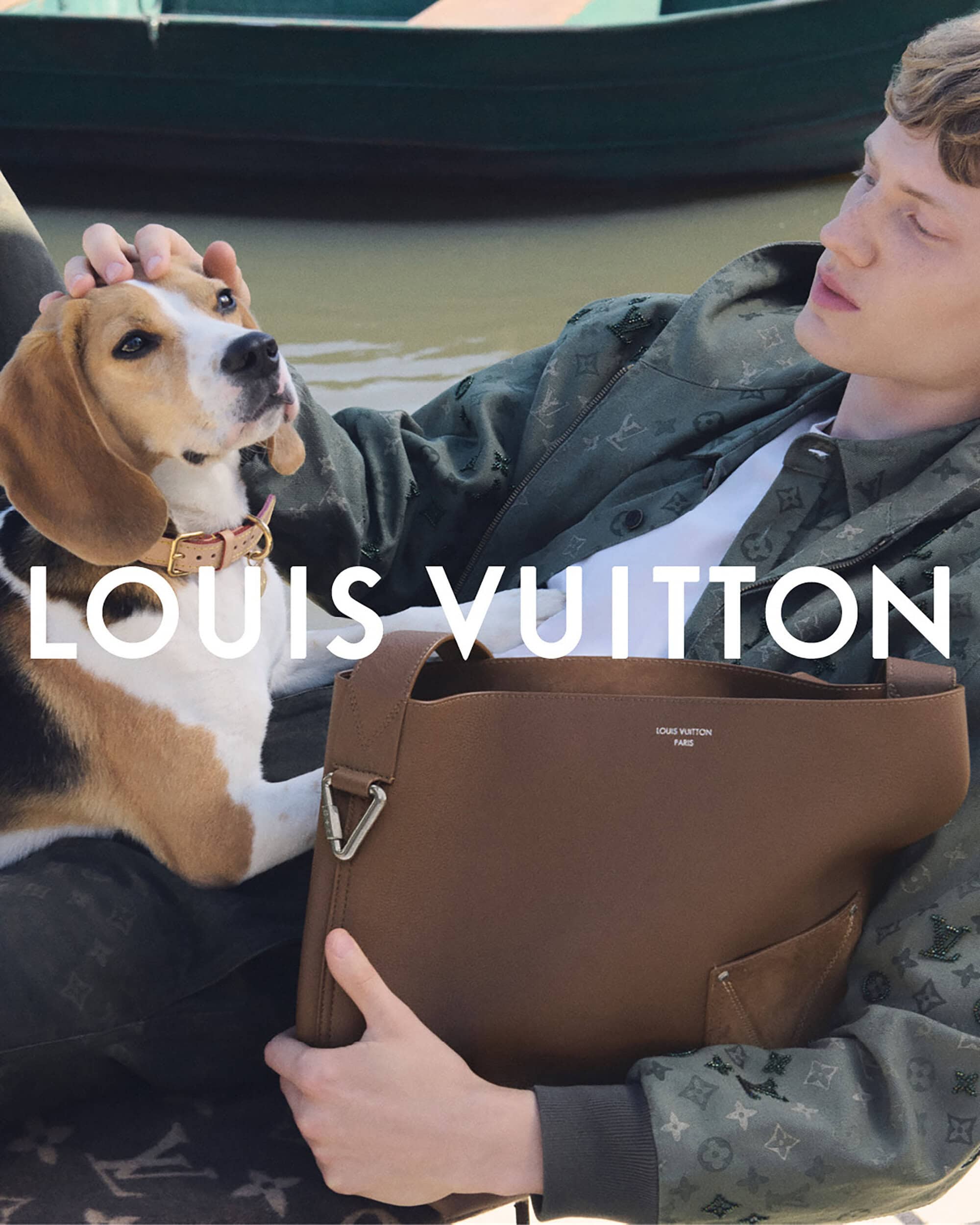 A light-skinned young adult male and a beagle dog pose with a Louis Vuitton purse outdoors, with the man petting the dog's head.