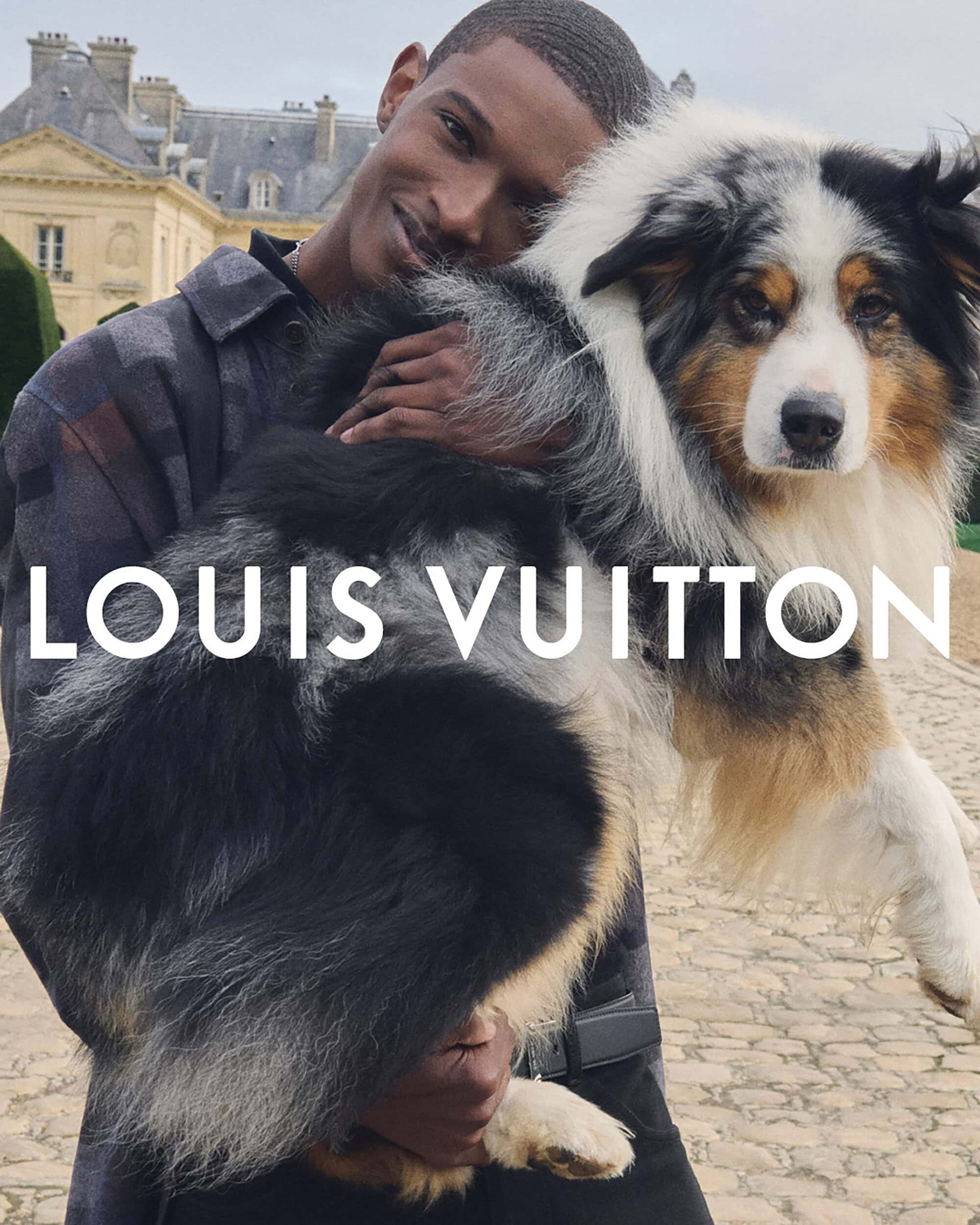 A man poses outdoors while holding an Australian Shepherd, showcasing a fashion collaboration with a historic building in the background.