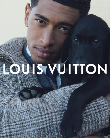 A black Labrador puppy is cradled by an adult male with short dark hair in a promotional image with the 'Louis Vuitton' logo.
