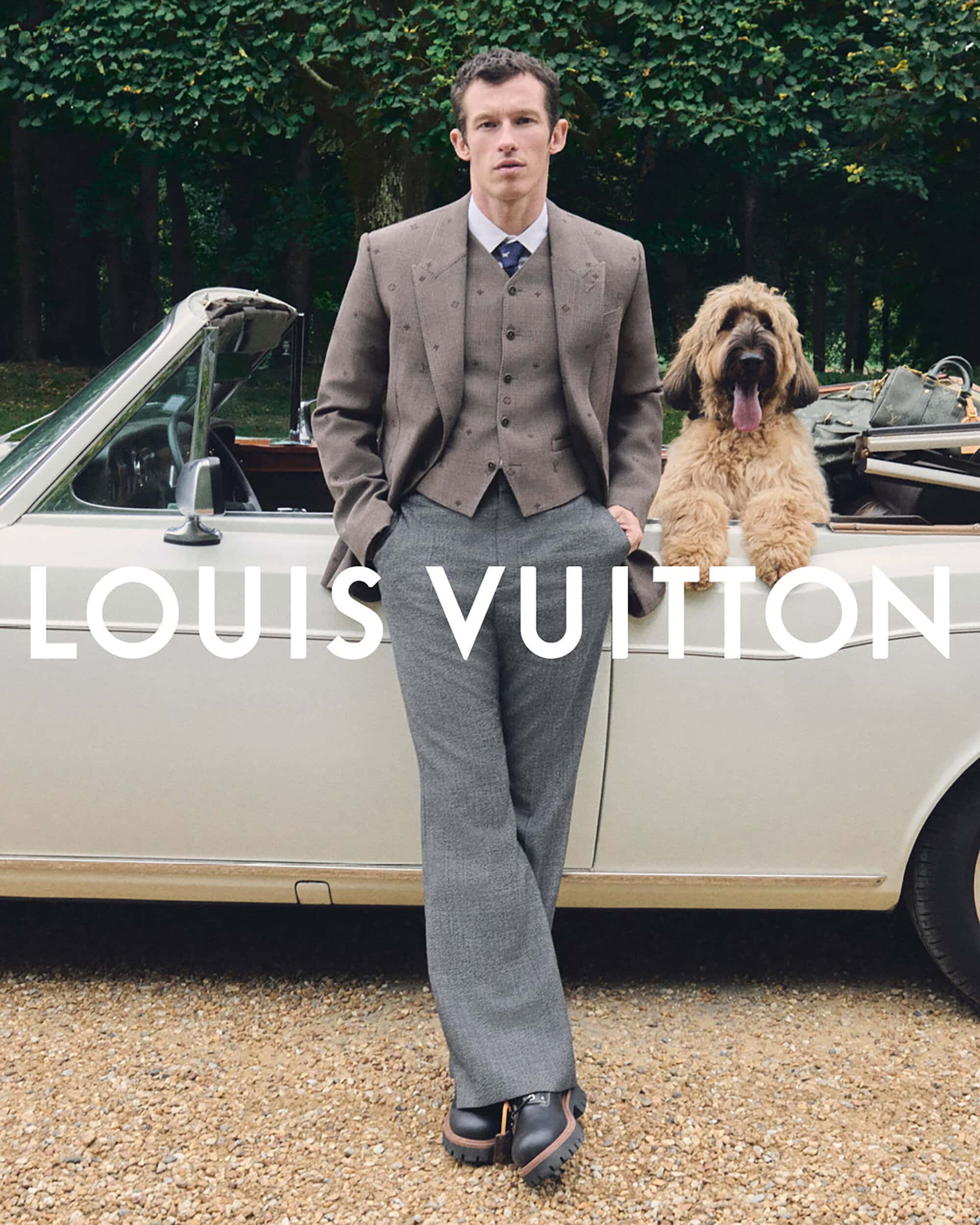 A stylish adult man and a Briard dog near a vintage convertible car against a backdrop of lush green trees. Outdoor setting, fashion.