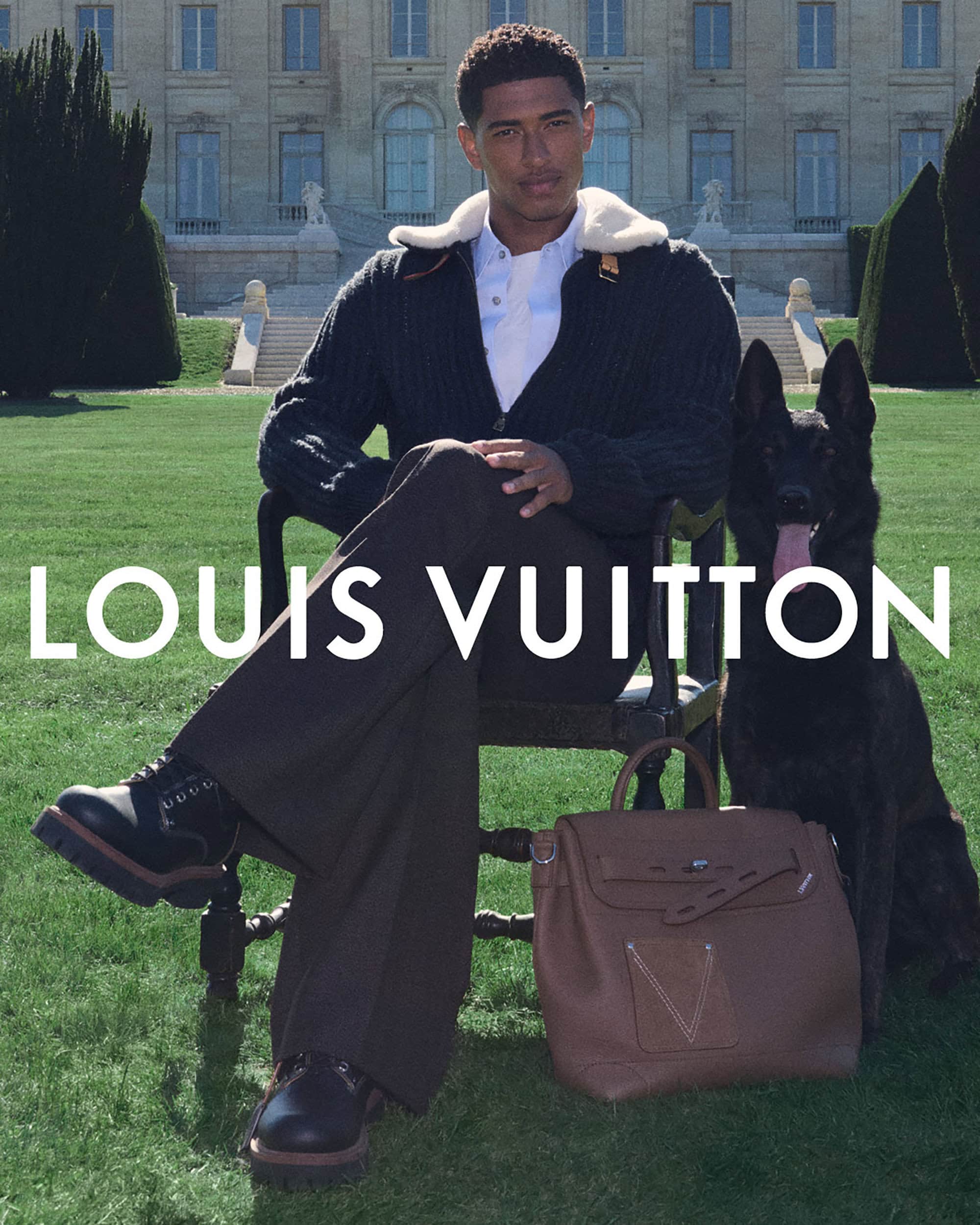 An adult male model sits on a chair next to a black dog, showcasing fashion and accessories in front of a grand building.