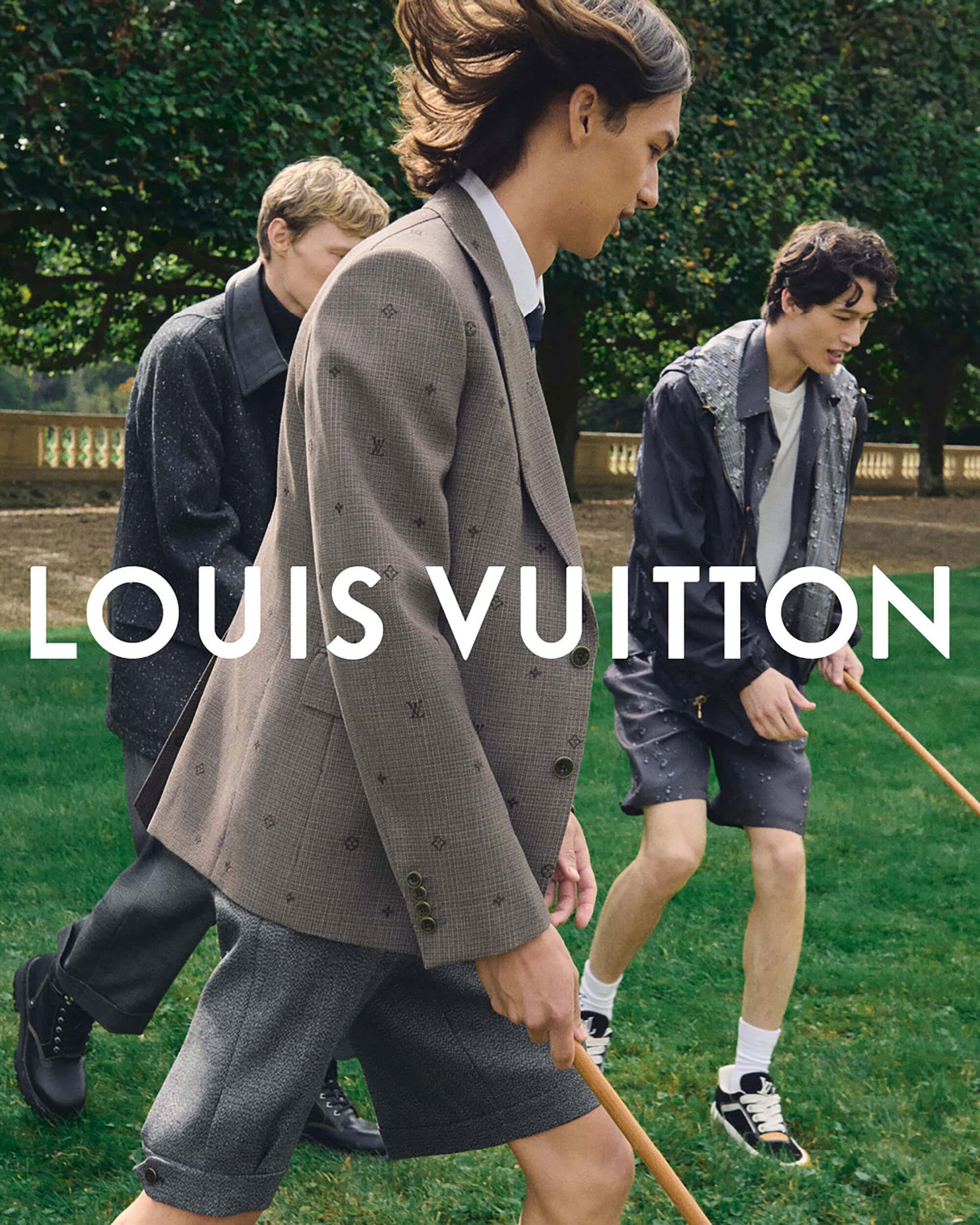 Three men in Louis Vuitton clothing playing lawn games outdoors on a green field with trees and a building in the background.