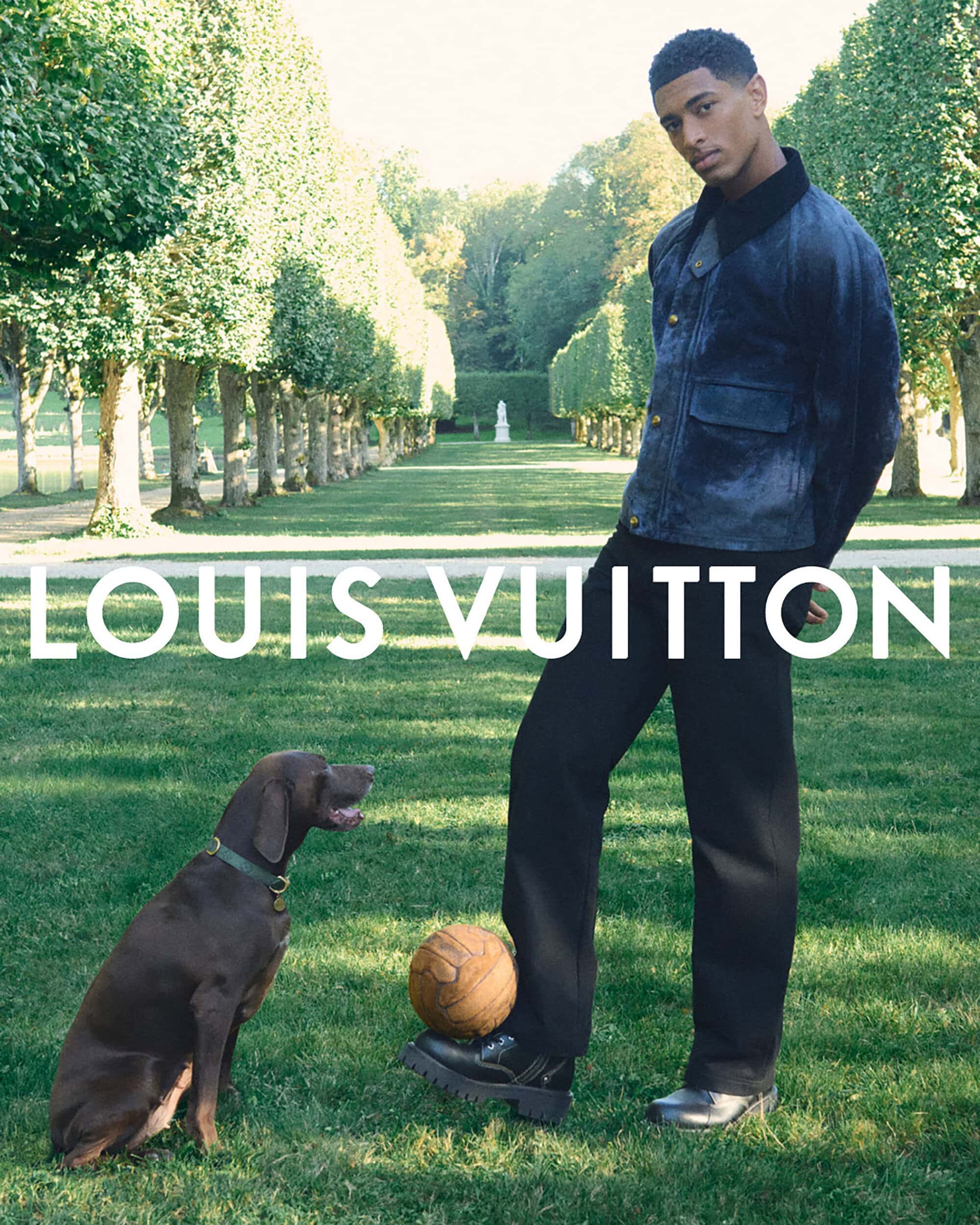 A young man in a jacket and trousers stands near a soccer ball, next to a brown dog, in a verdant green field at daytime for Louis Vuitton.