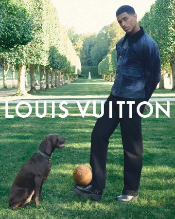 A young man in a jacket and trousers stands near a soccer ball, next to a brown dog, in a verdant green field at daytime for Louis Vuitton.