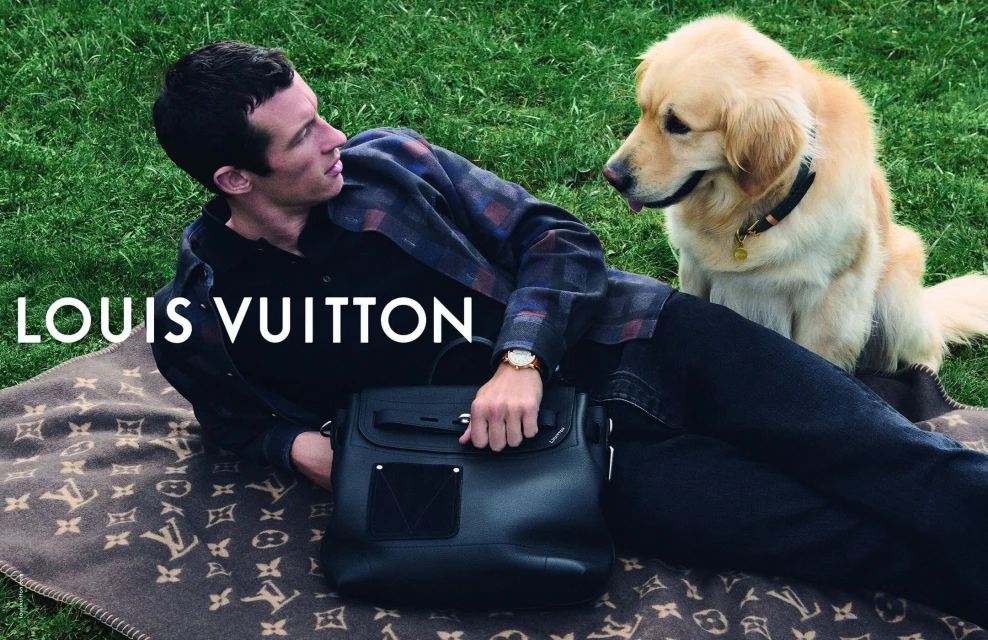 Adult male and Golden Retriever dog pose with Louis Vuitton blanket and bag on green lawn in outdoor setting for fashion advertisement.