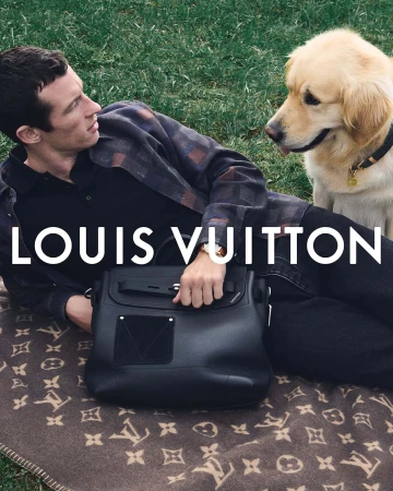 Man reclining on a monogrammed blanket next to a golden retriever, holding a large black leather bag.
