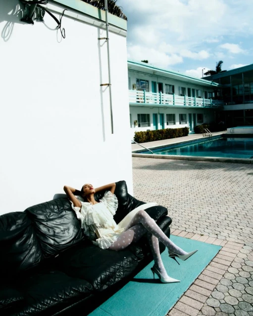 Woman in white dress relaxes on black leather couch near swimming pool, bathed in bright sunlight against a blue sky with clouds.
