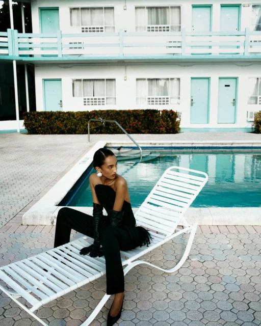 Woman in black outfit sitting on white lounge chair next to a pool with a motel in the background on a sunny day.