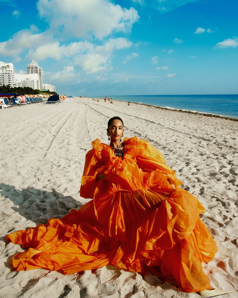 A model poses on a sandy beach in a voluminous, bright orange gown, with city buildings and blue sky with clouds in the distance.