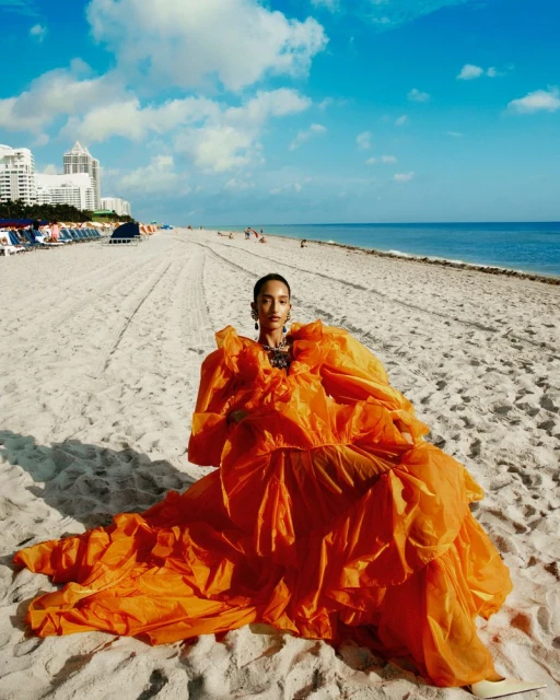 A model poses on a sandy beach in a voluminous, bright orange gown, with city buildings and blue sky with clouds in the distance.