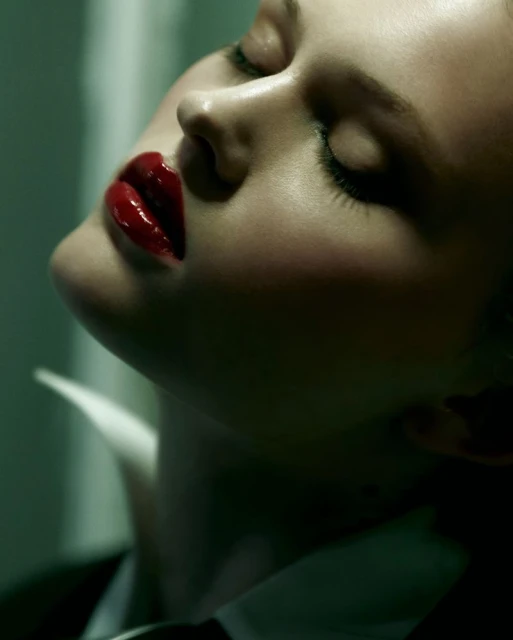 Close-up of a fair-skinned woman with closed eyes, glossy red lips, wearing a black suit jacket and white collared shirt, tilting her head back.