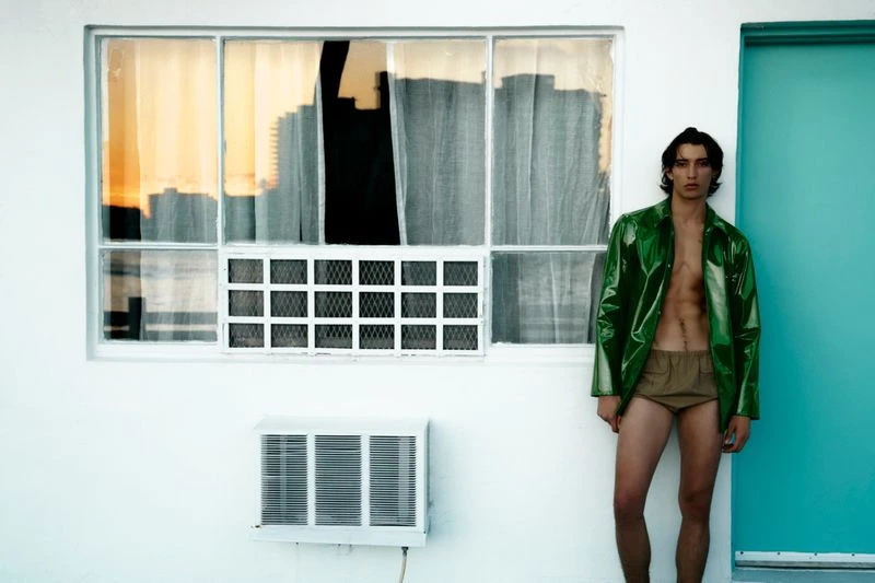 A male model poses in front of a white wall wearing a translucent green jacket and khaki shorts; cityscape reflections in a window.