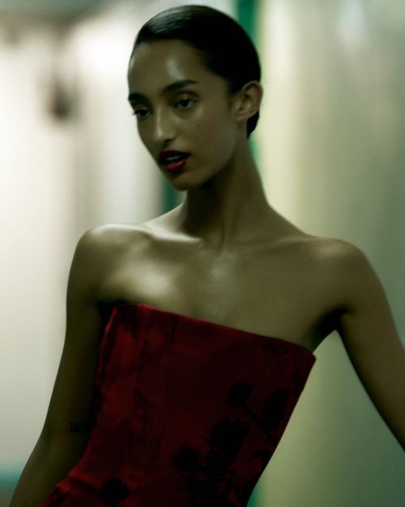 Portrait of a woman with dark hair and red lipstick wearing a red strapless dress in front of a neutral background. Fashion photography.