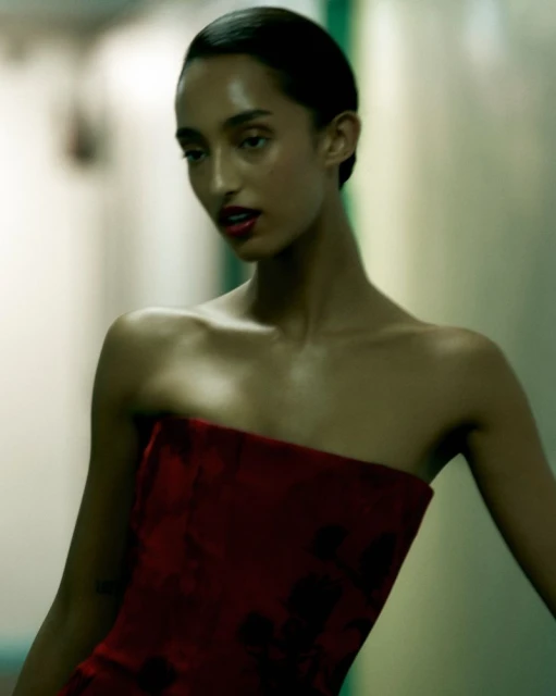 Portrait of a woman with dark hair and red lipstick wearing a red strapless dress in front of a neutral background. Fashion photography.
